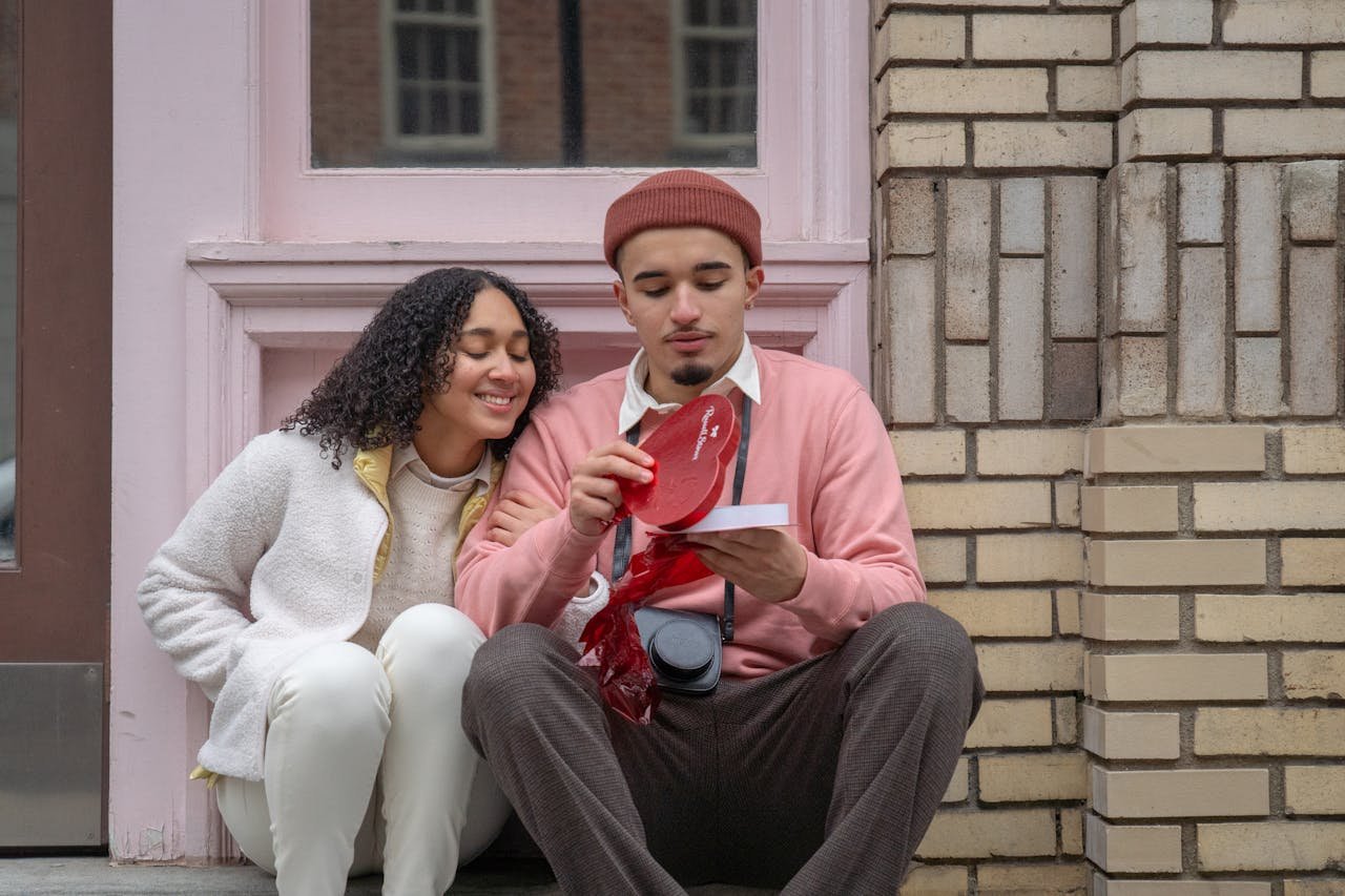 Young ethnic guy in stylish outfit opening heart shaped box with chocolate while sitting on street with happy excited girlfriend on Saint Valentines Day